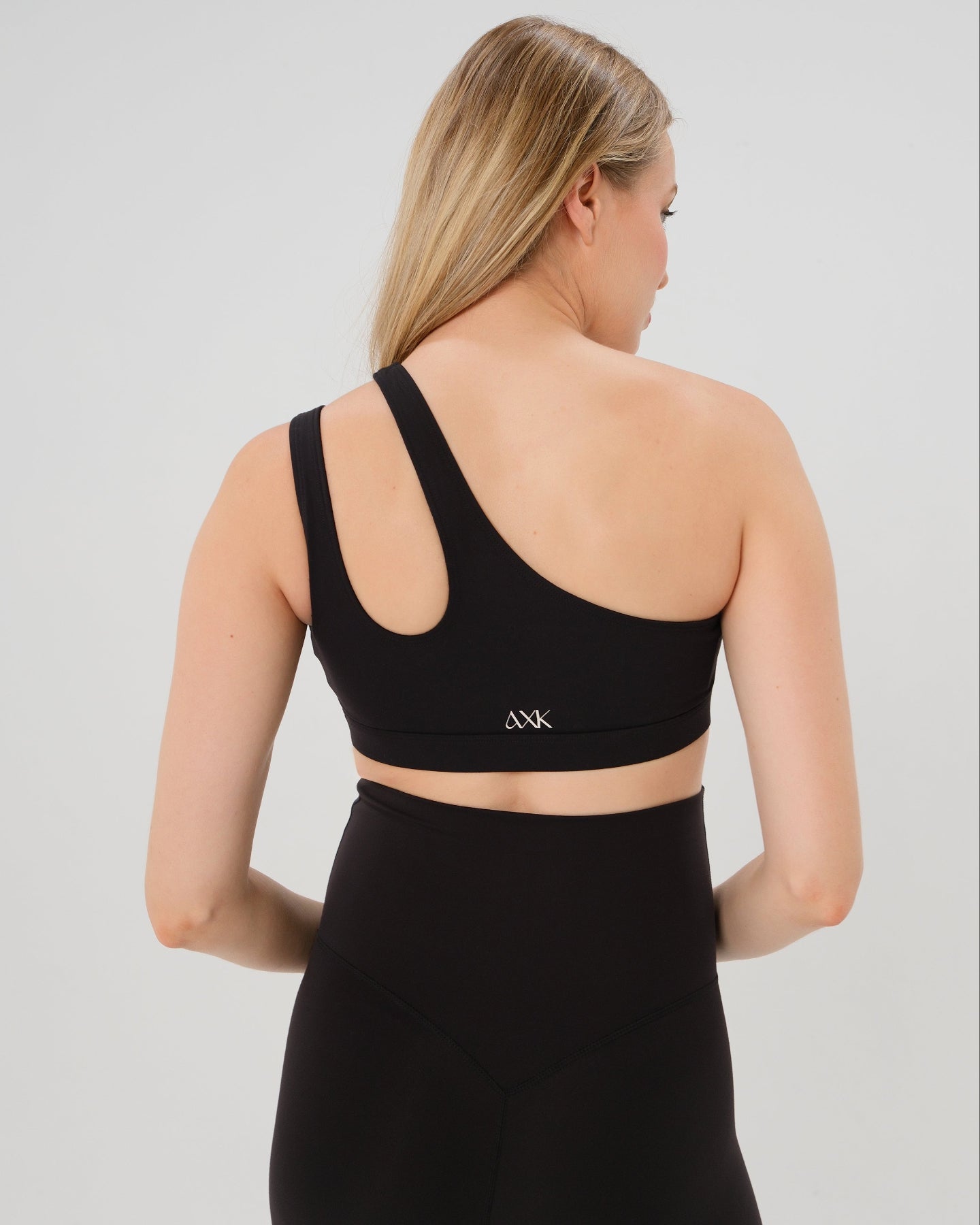 Pregnant woman in a one-shoulder black maternity and nursing sports bra, wearing matching black maternity leggings. Best selling 2026 premium maternity athletic and activewear by AXK, offered in regular and plus sizes, with glowing 5 star reviews.