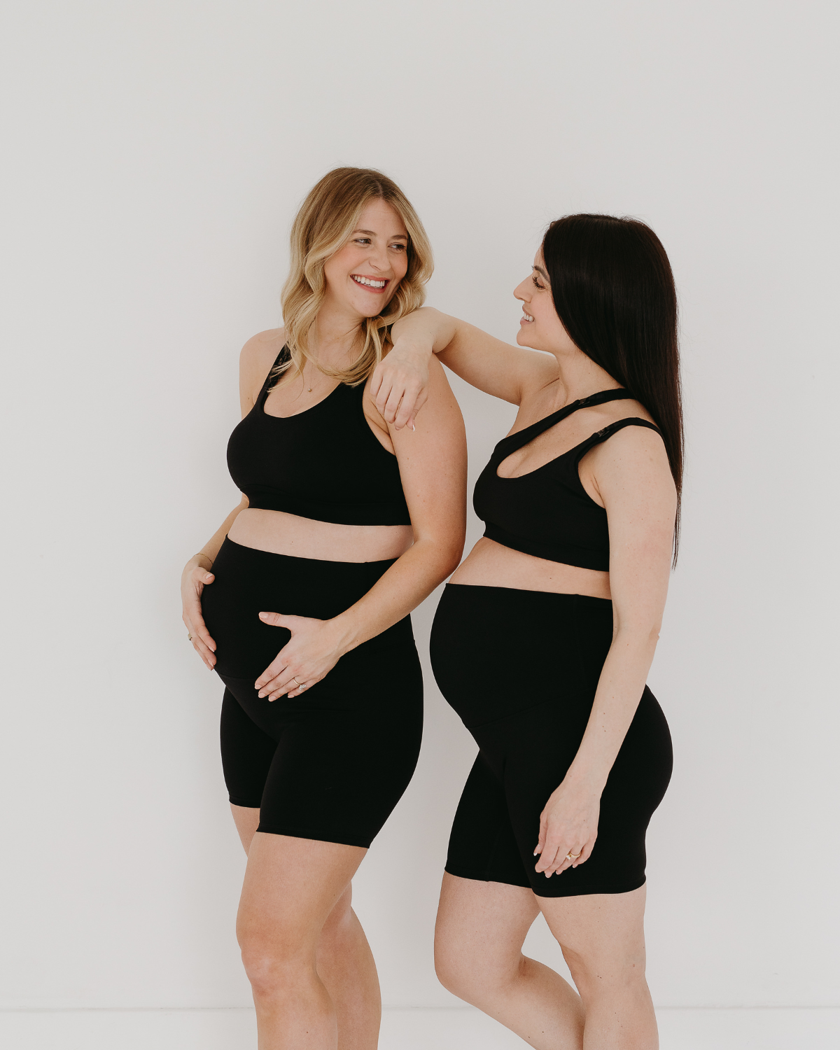 Two pregnant women wearing premium black maternity bike shorts with supportive high waist. One is wearing a one-shoulder, low-impact, nursing bra and one is wearing a matching high-impact, racerback maternity and nursing sports bra. They are smiling at each other and standing together against a white background. Premium maternity athletic and activewear by AXK.