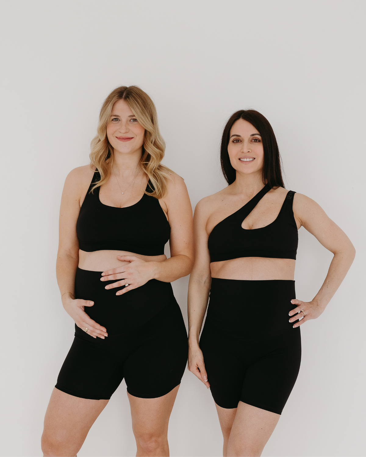 Two pregnant women wearing premium black maternity bike shorts with supportive high waist. One is wearing a one-shoulder, low-impact, nursing bra and one is wearing a matching high-impact, racerback maternity and nursing sports bra. They are standing together against a white background. Premium maternity athletic and activewear by AXK.