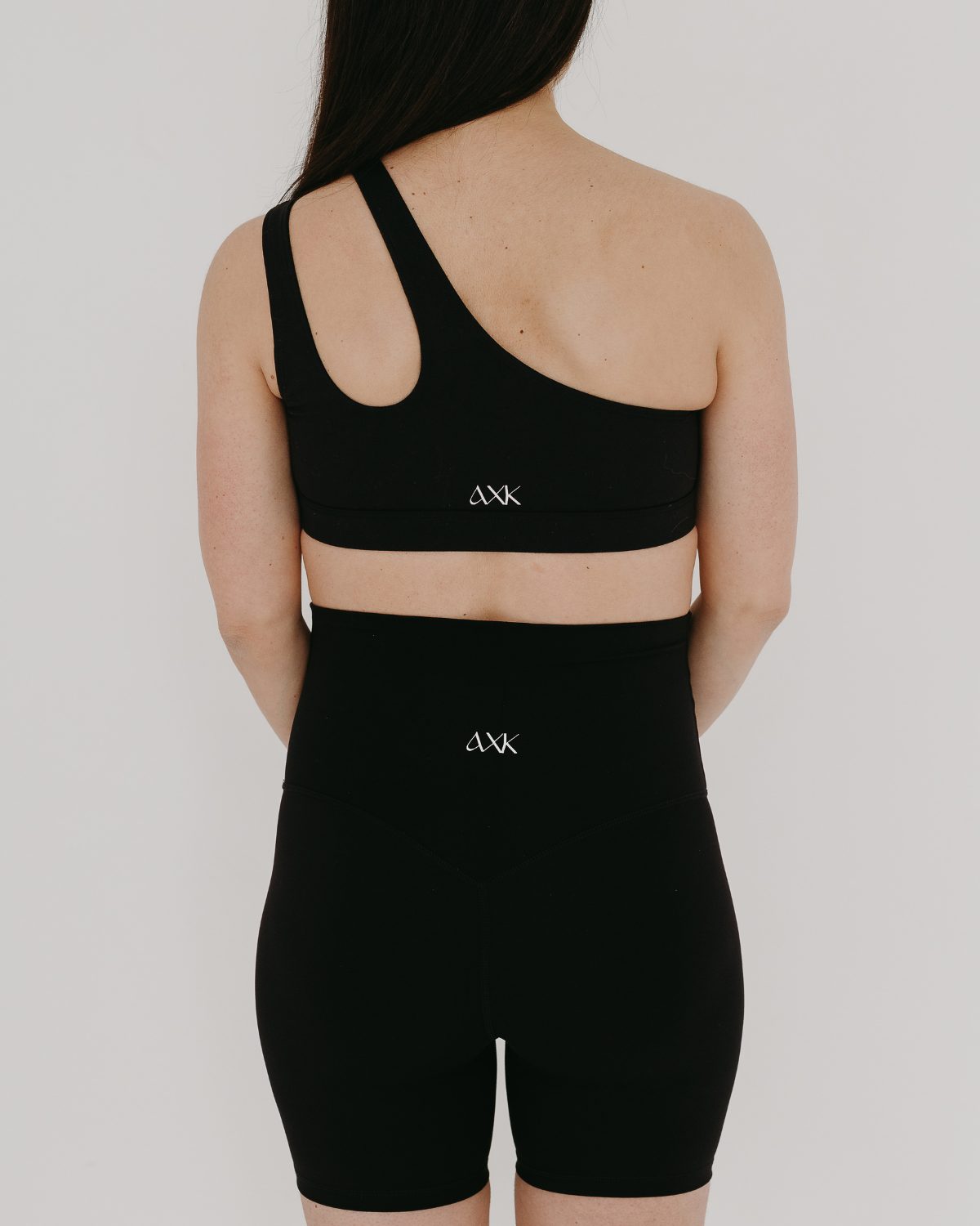 Back view of woman wearing premium black maternity bike shorts and a matching one-shoulder maternity and nursing sports bra, both featuring the AXK logo, standing against a white background. Premium maternity athletic and activewear by AXK.