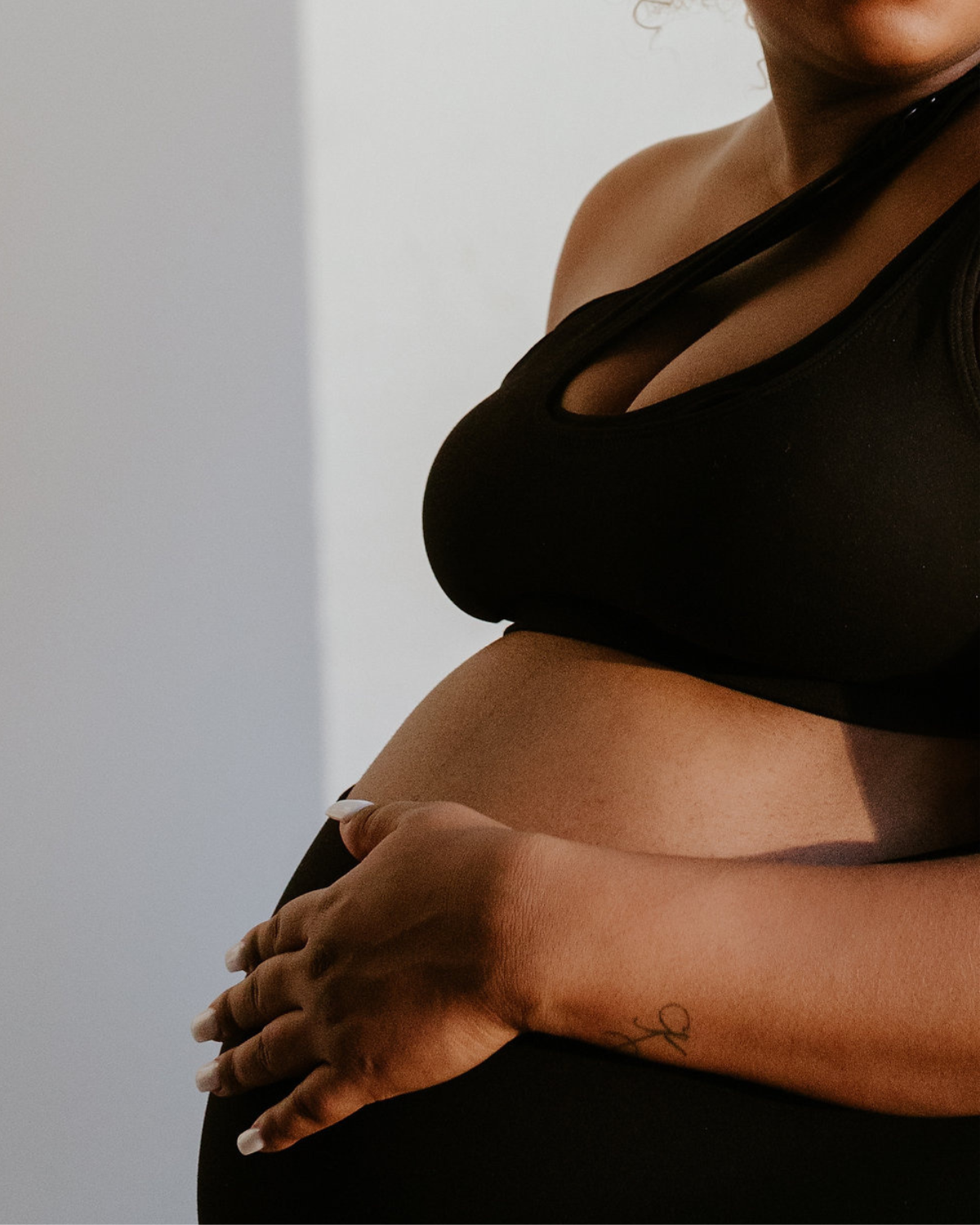 Close-up of a pregnant woman wearing a premium black one-shoulder, low-impact maternity and nursing sports bra, cradling her belly. She is standing in front of a white wall with soft natural light. Premium maternity activewear by AXK.