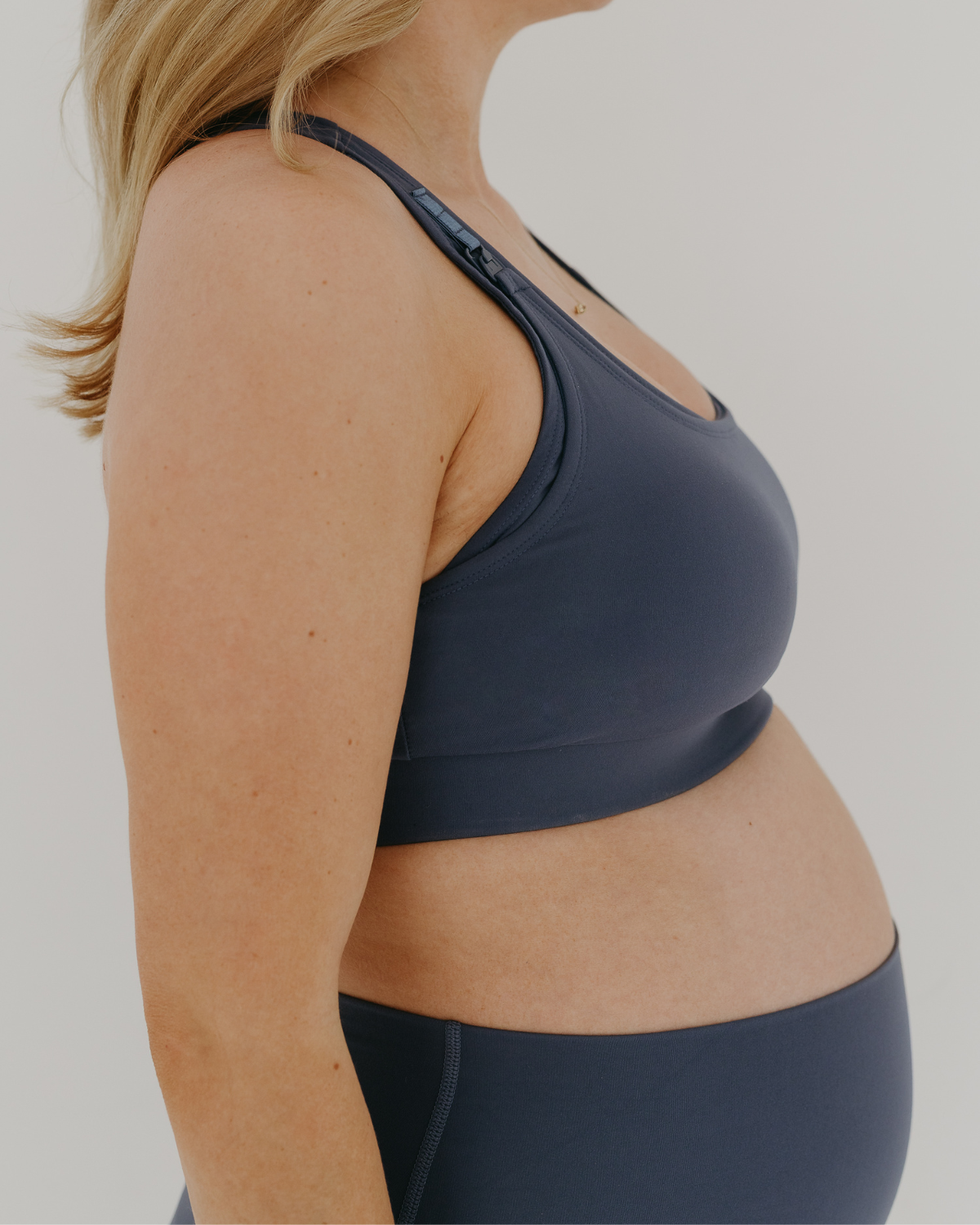 Close up side view of a blue high-impact, racerback, maternity and nursing sports bra with one-handed clip access. Worn by a pregnant model, standing in front of a white background. Premium maternity athletic and activewear by AXK.