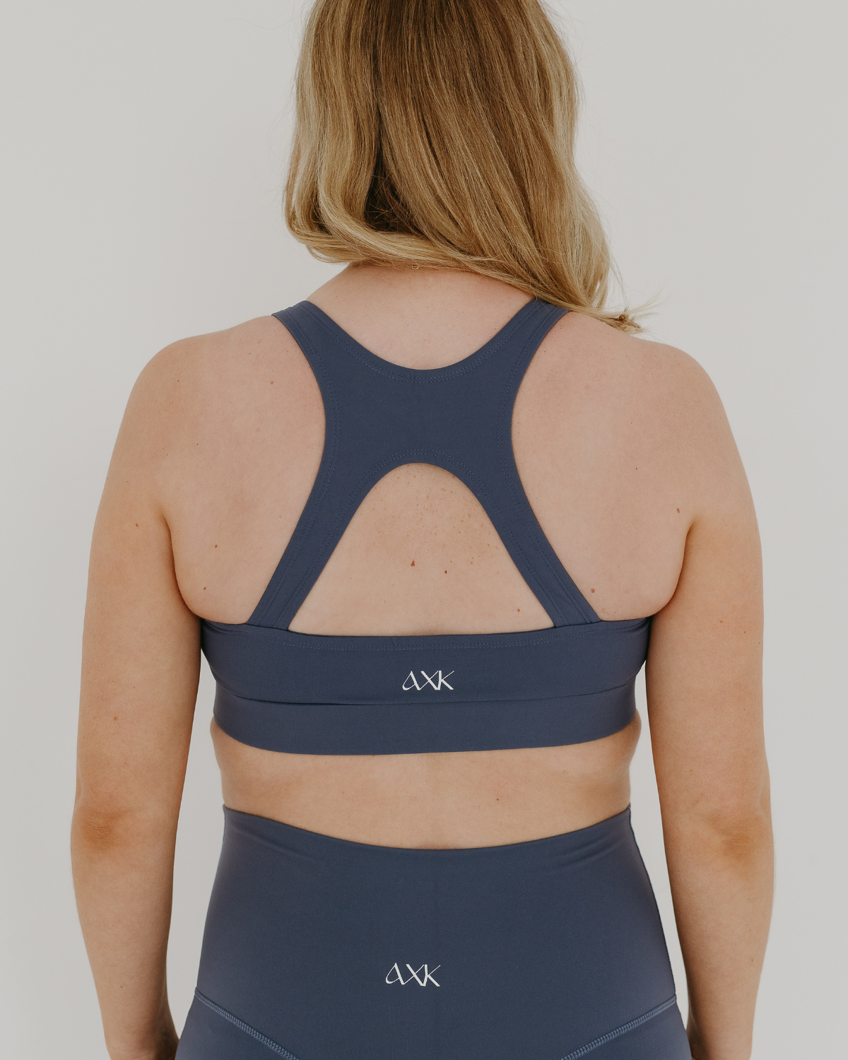 Back view of a blue high-impact, racerback, maternity and nursing sports bra with one-handed clip access. Worn by a pregnant model, standing in front of a white background. Premium maternity athletic and activewear by AXK.