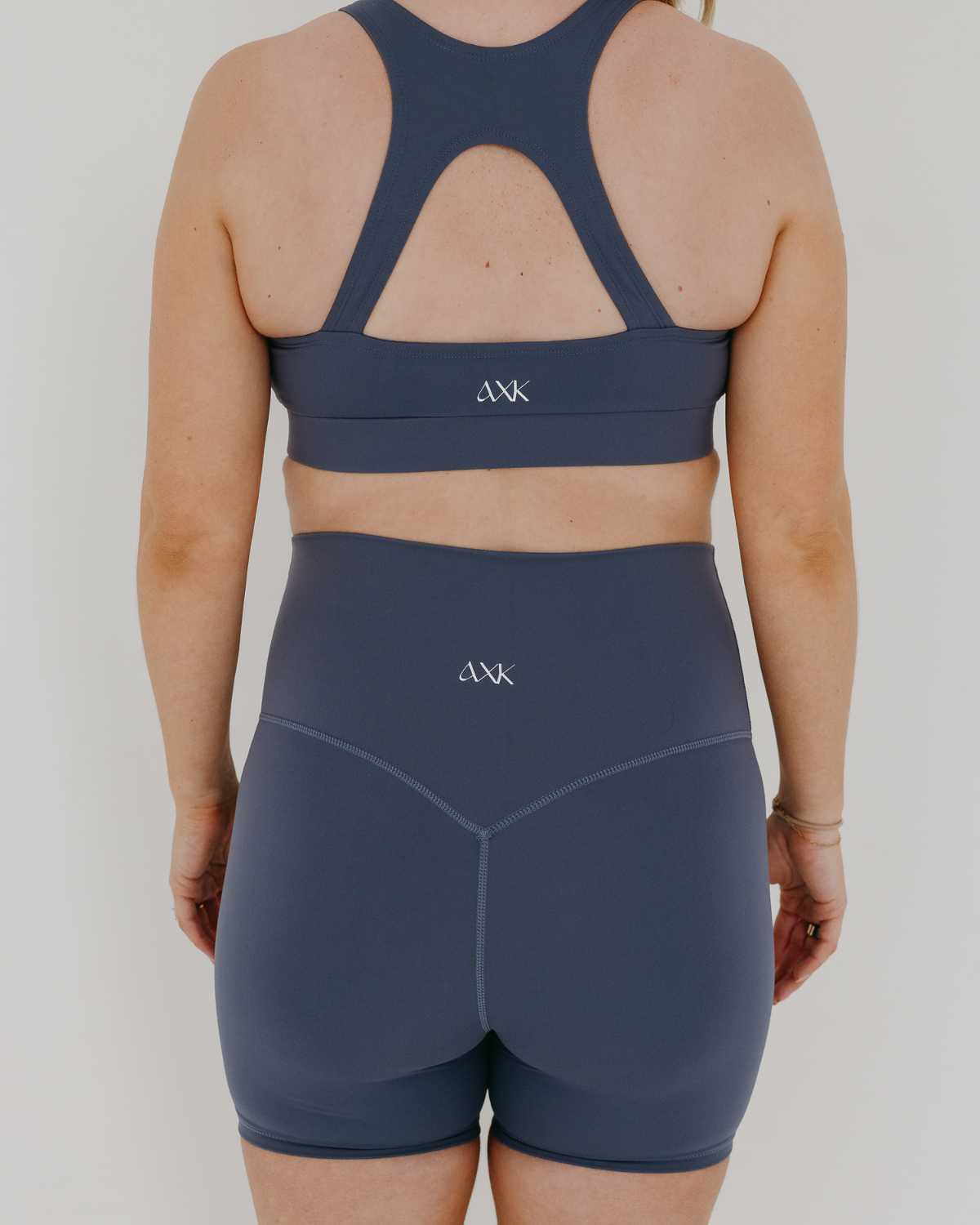 Pregnant woman wearing our premium blue best reviewed maternity bike shorts for 2026 with a supportive high waist and matching high-impact, racerback maternity and nursing sports bra, standing against a white background. Premium maternity athletic and activewear by AXK.