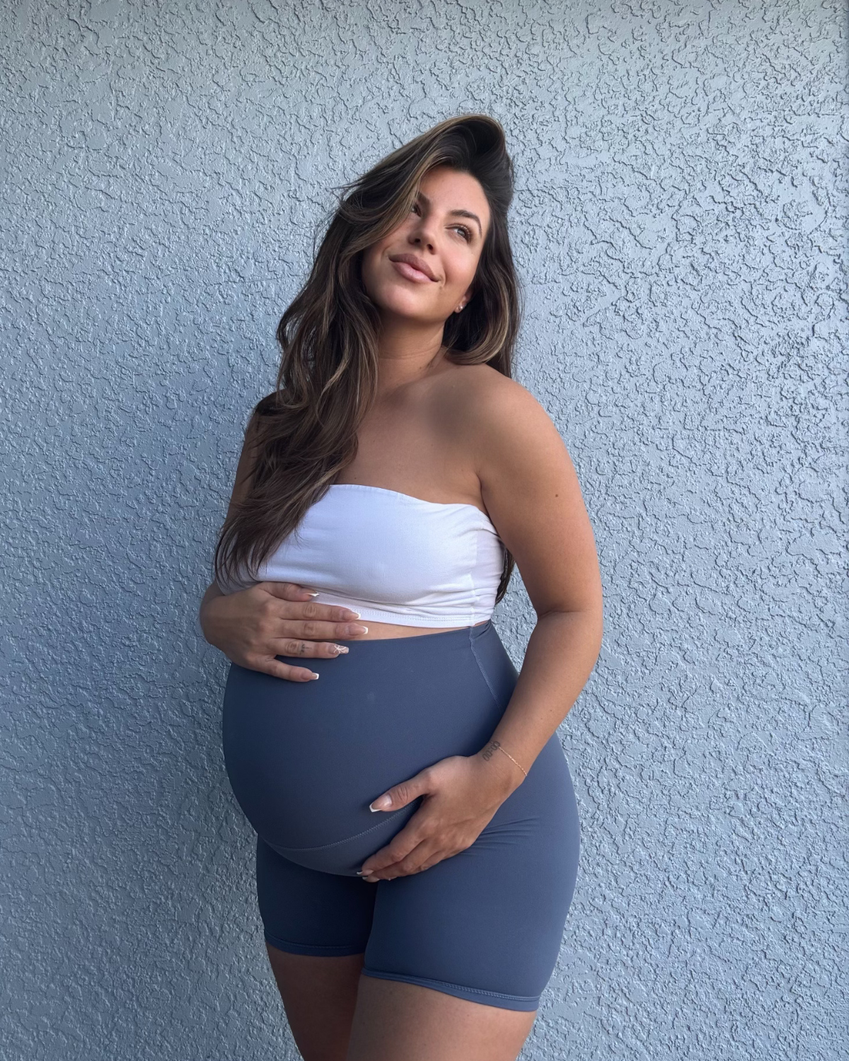 Pregnant woman wearing premium blue maternity bike shorts with a supportive high waist, paired with a white bandeau top, standing outdoors against a textured light blue wall. Premium maternity athletic and activewear by AXK.