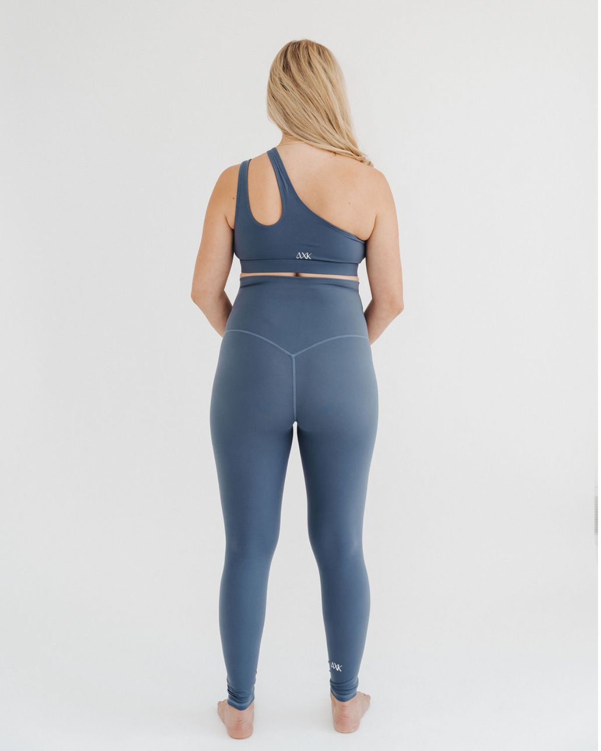 Back view of pregnant woman wearing premium blue maternity leggings with a supportive high waist and V-seam back, paired with a matching one-shoulder maternity and nursing sports bra, standing against a white background. Premium maternity athletic and activewear by AXK.