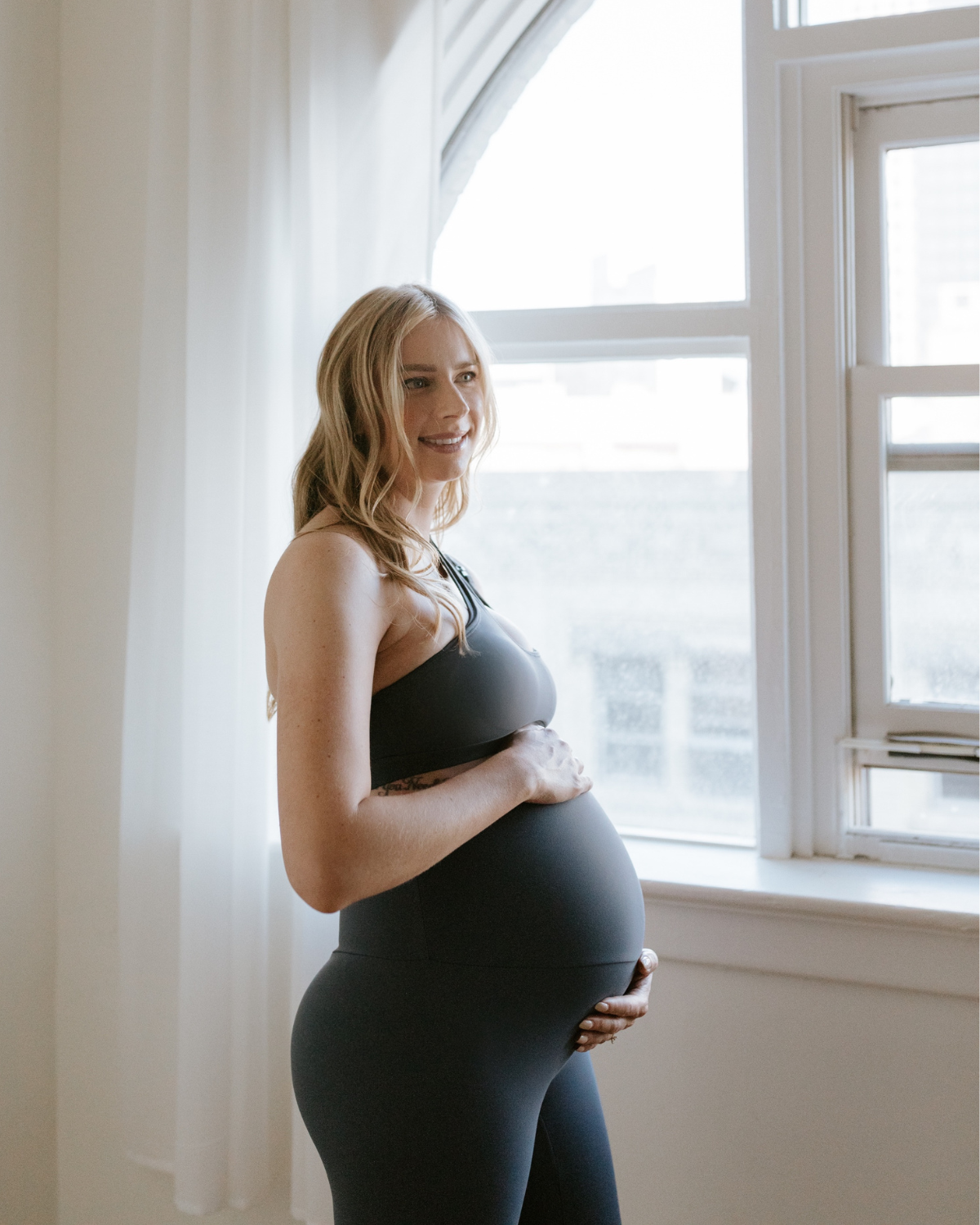 Pregnant woman wearing premium slate maternity leggings with a flattering scrunch butt and matching one-shoulder, nursing sports bra, smiling while standing near a bright window. Premium maternity athletic and activewear by AXK.