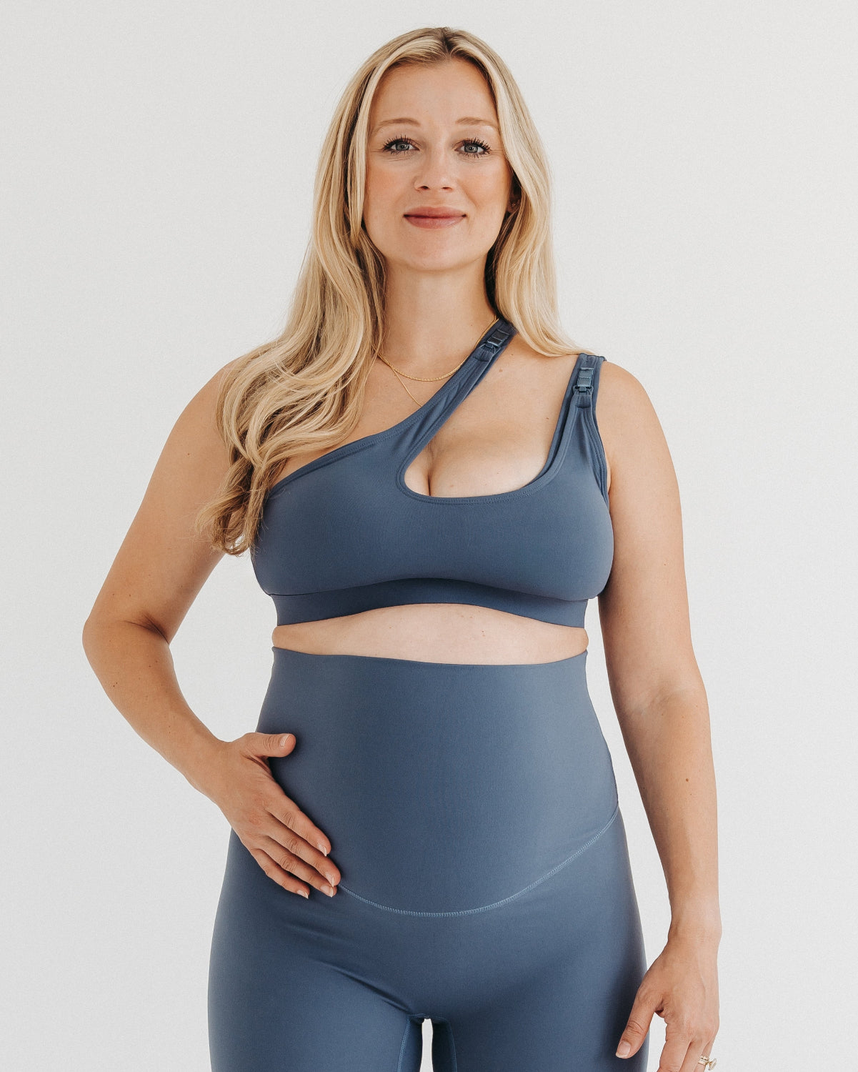 Pregnant woman wearing premium maternity leggings with a supportive high waist and a matching one-shoulder maternity and nursing sports bra in blue, standing against a white background. Premium maternity athletic and activewear by AXK in plus size, tall friendly, and petite friendly options.