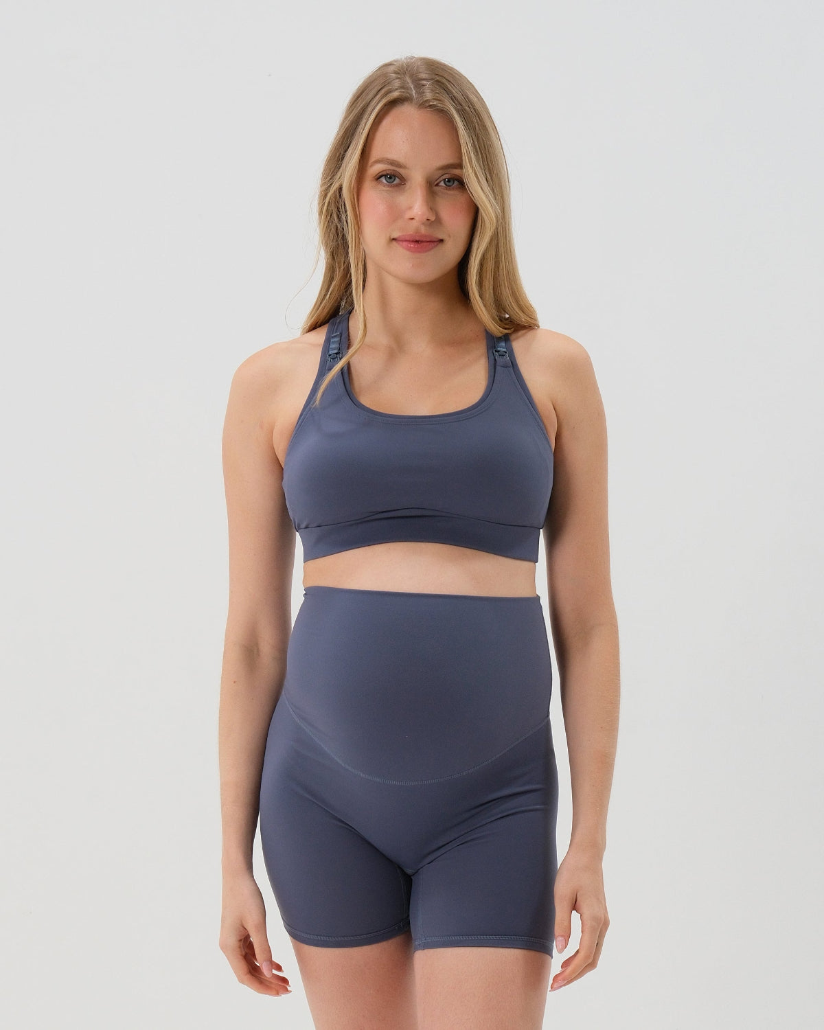 Pregnant woman wearing our premium blue best reviewed maternity bike shorts for 2026 with a supportive high waist and matching high-impact, racerback maternity and nursing sports bra, standing against a white background. Premium maternity athletic and activewear by AXK.
