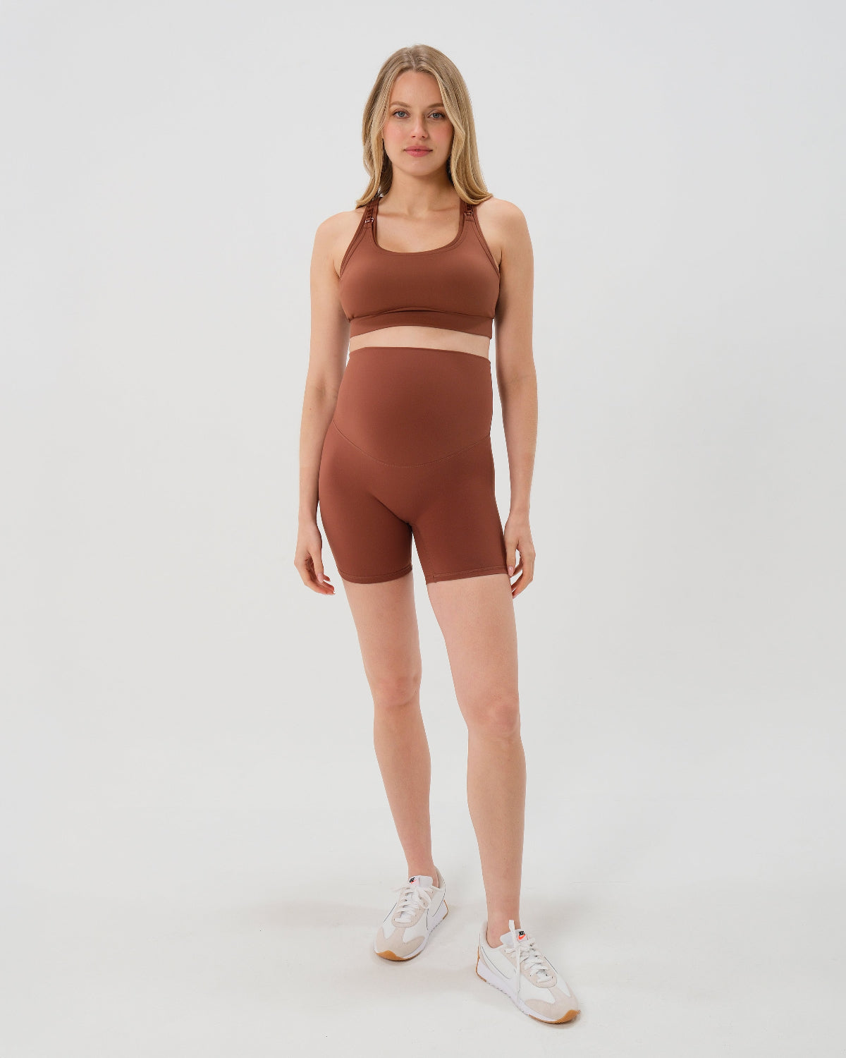 Pregnant woman wearing our best reviewed premium brown maternity bike shorts with a supportive high waist and matching high-impact, racerback maternity and nursing sports bra, standing against a white background. Premium maternity athletic and activewear by AXK - high quality petite, tall, and plus size maternity.