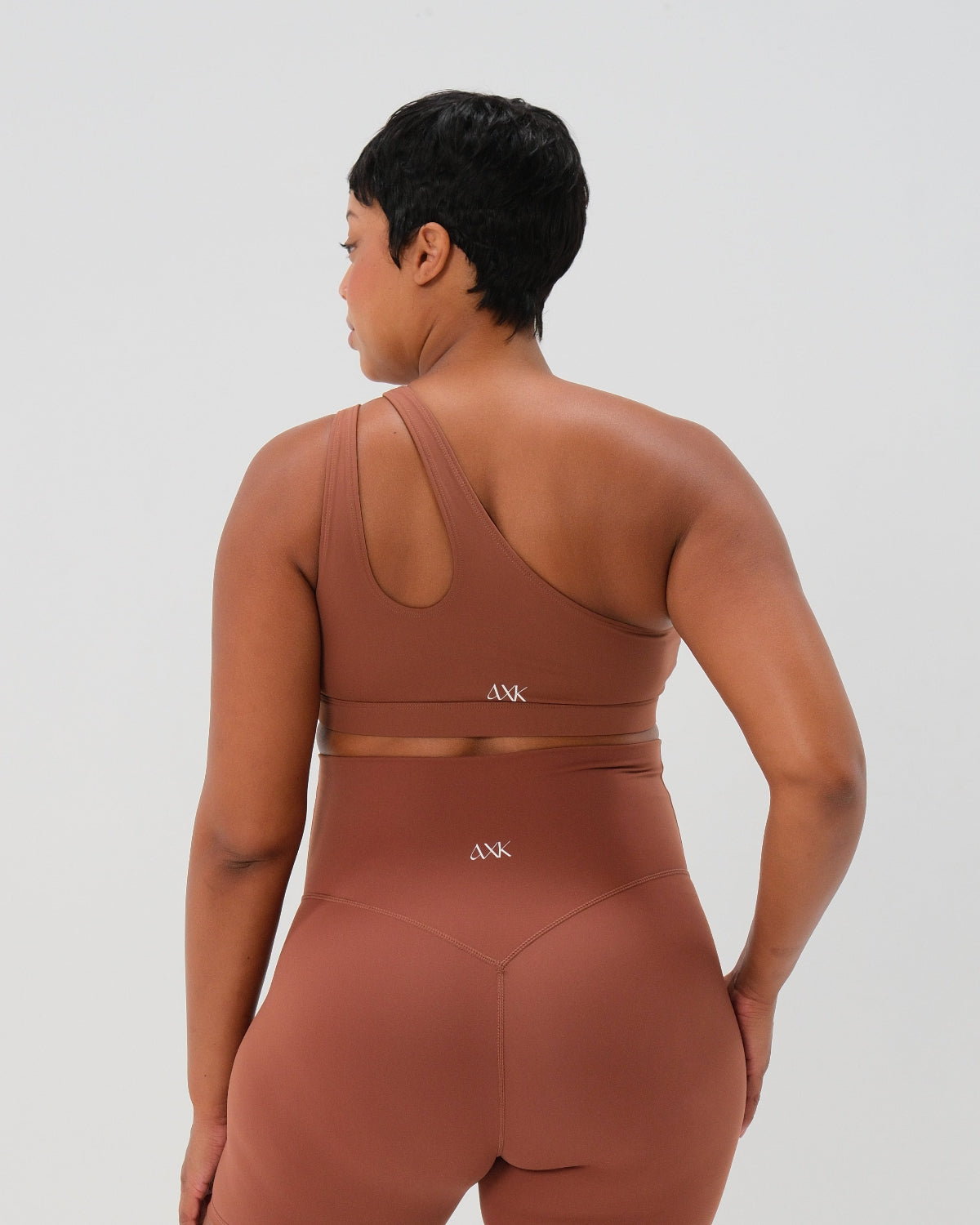 Back view of brown one-shoulder maternity and nursing sports bra and matching leggings. The bra features one-handed clip access for easy breastfeeding. Premium maternity activewear by AXK is offered in petite and plus size.