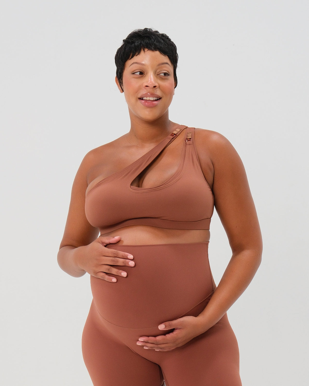 Close-up of a pregnant woman wearing a brown one-shoulder, maternity and nursing sports bra and leggings, with one hand resting on her baby bump. Premium maternity activewear by AXK.