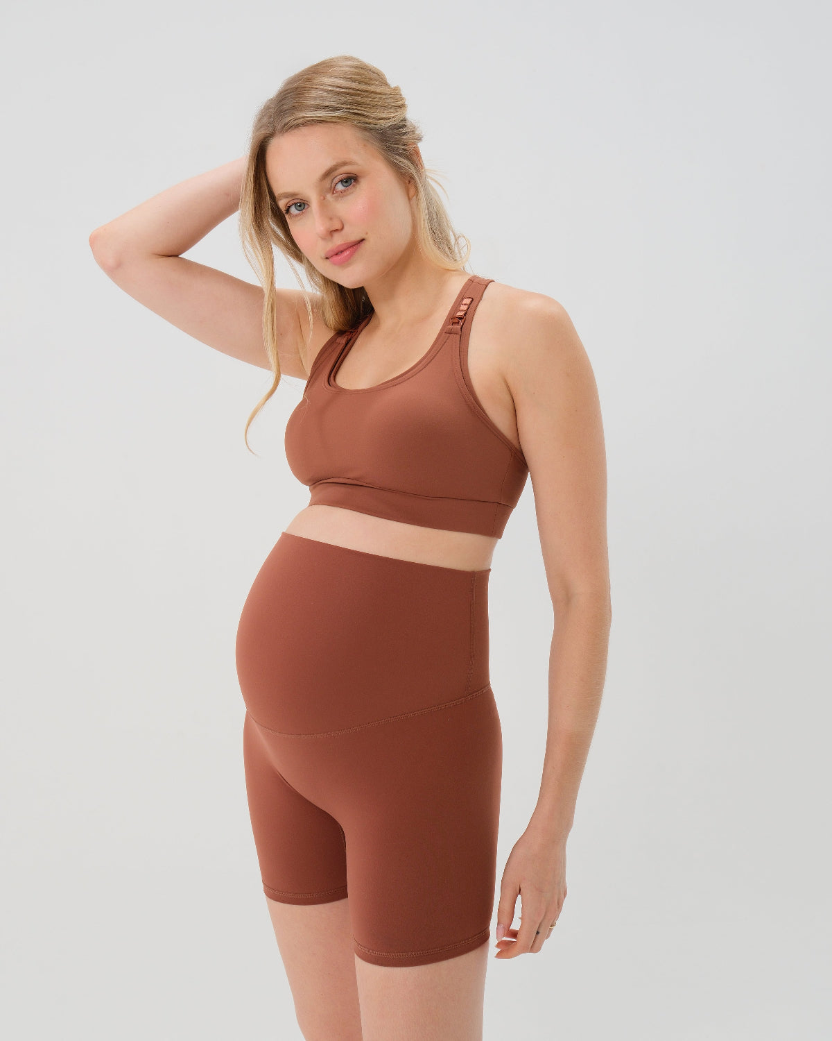 Pregnant woman wearing our best reviewed premium brown maternity bike shorts with a supportive high waist and matching high-impact, racerback maternity and nursing sports bra, standing against a white background. Premium maternity athletic and activewear by AXK - high quality petite, tall, and plus size maternity.