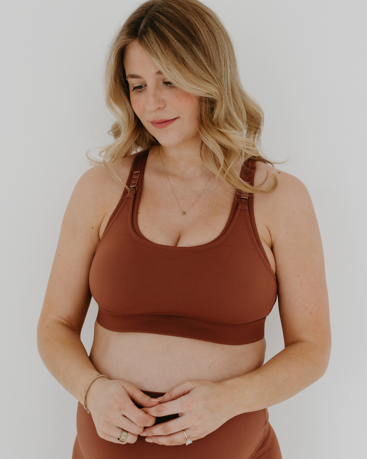 Close up view of a pregnant woman wearing a brown high-impact, racerback, maternity and nursing sports bra and matching high-waist maternity leggings. She has her hands cradling her belly standing in front of a white background. Premium maternity athletic and activewear by AXK.