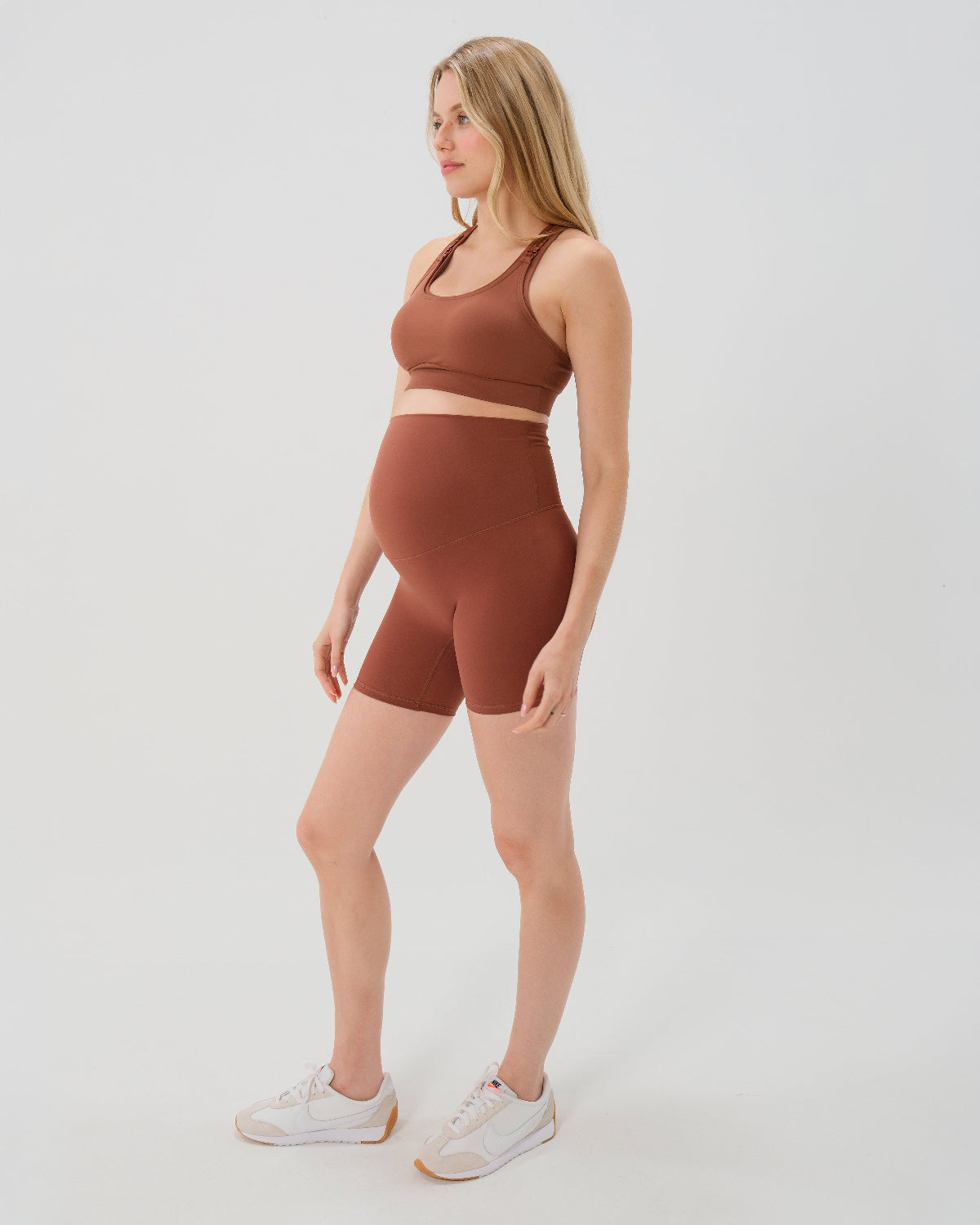 Pregnant woman wearing our best reviewed premium brown maternity bike shorts with a supportive high waist and matching high-impact, racerback maternity and nursing sports bra, standing against a white background. Premium maternity athletic and activewear by AXK - high quality petite, tall, and plus size maternity.