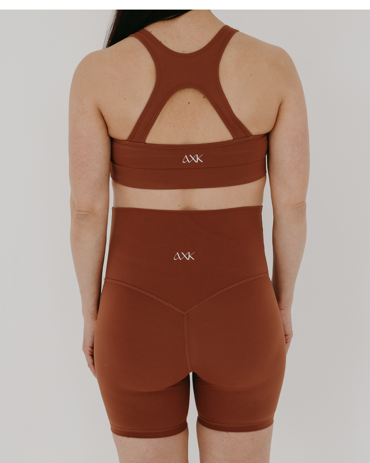 Back view of woman wearing premium brown maternity bike shorts with a supportive high waist and matching high-impact, racerback maternity and nursing sports bra, both featuring the AXK logo, standing against a white background. Premium maternity athletic and activewear by AXK.