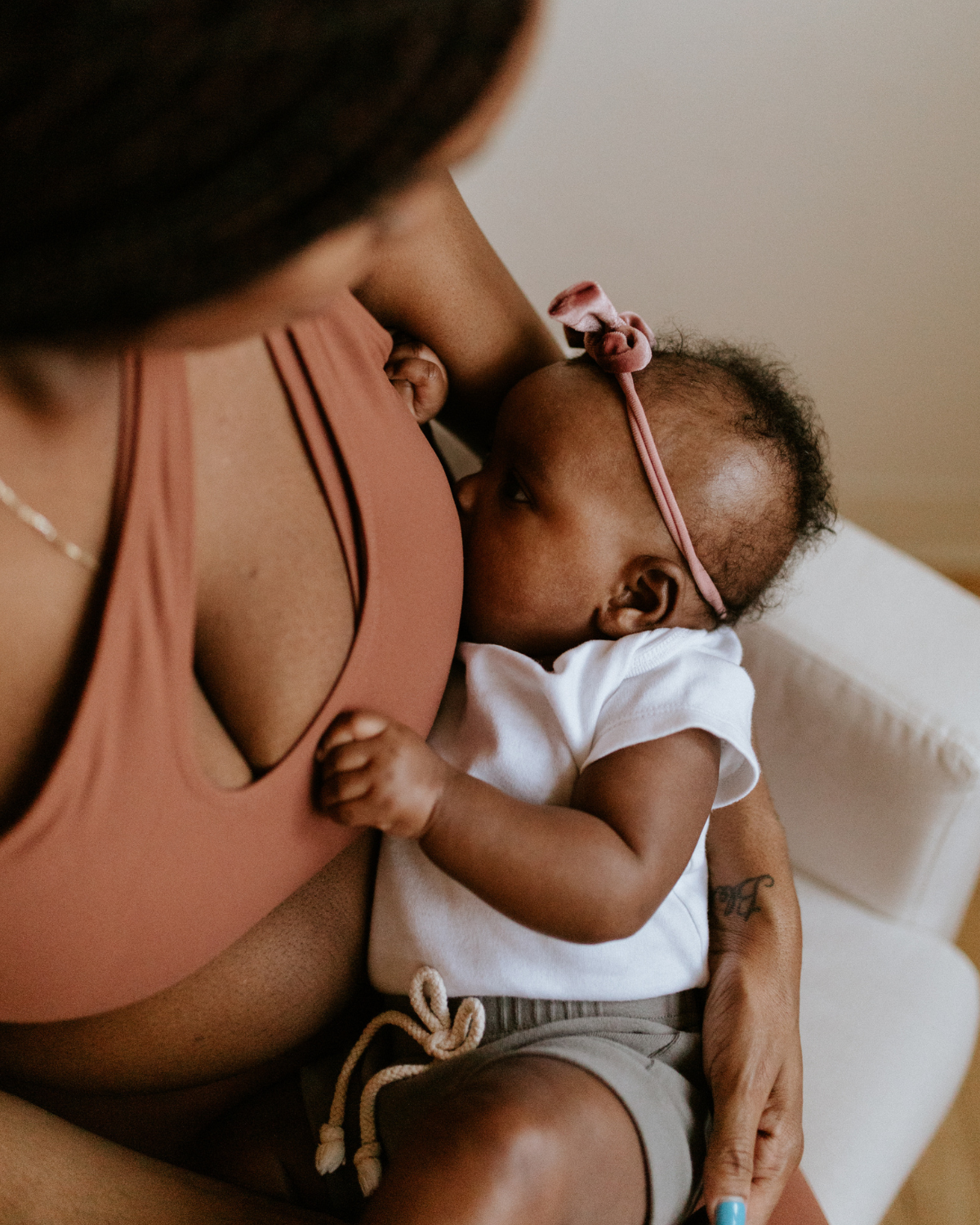 Mother holding her baby while wearing a brown one-shoulder, maternity and nursing sports bra, while seated indoors. AXK premium maternity activewear.