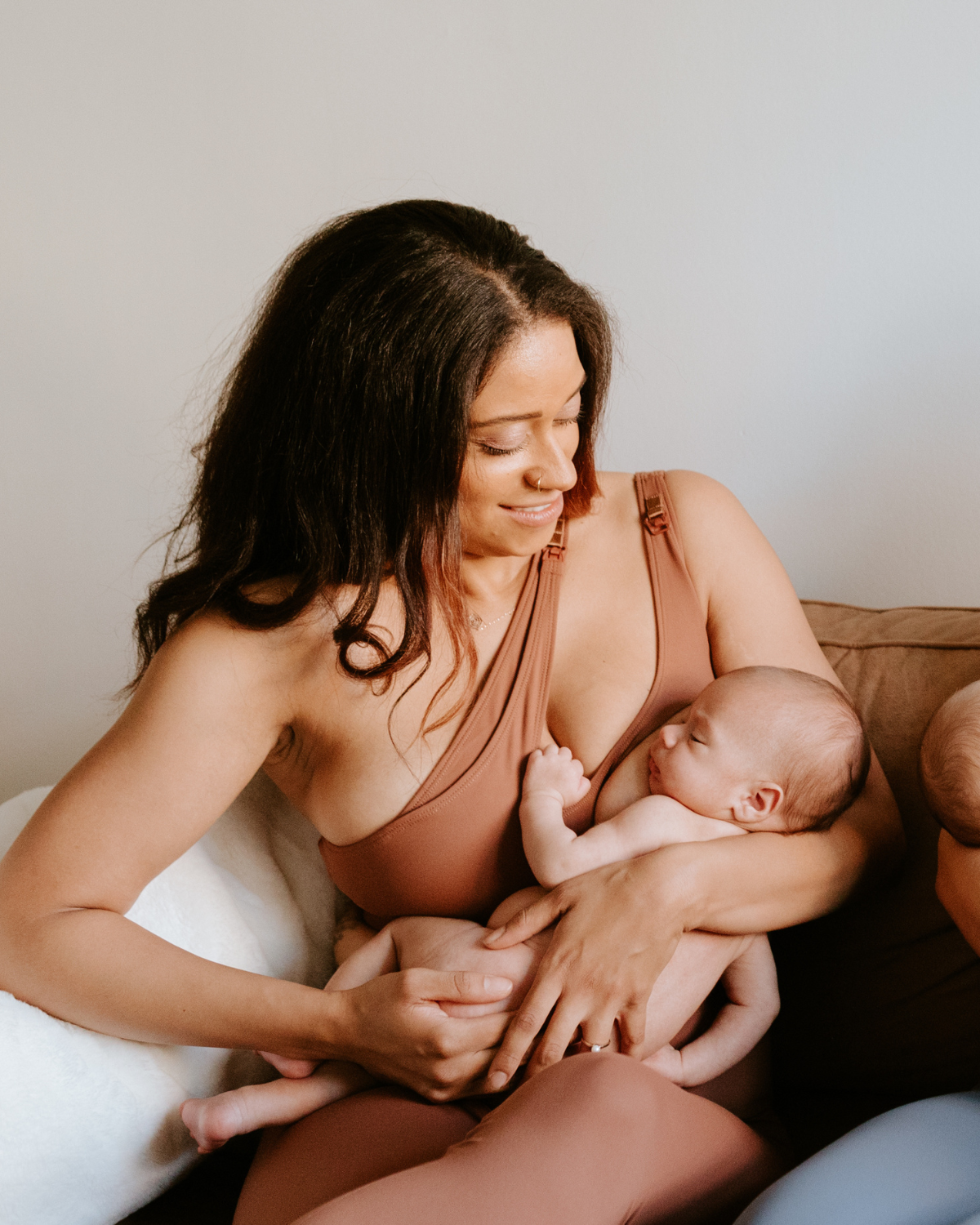 Mother wearing a brown one-shoulder, maternity and nursing sports bra and matching leggings, while holding and nursing a newborn baby. AXK premium maternity activewear