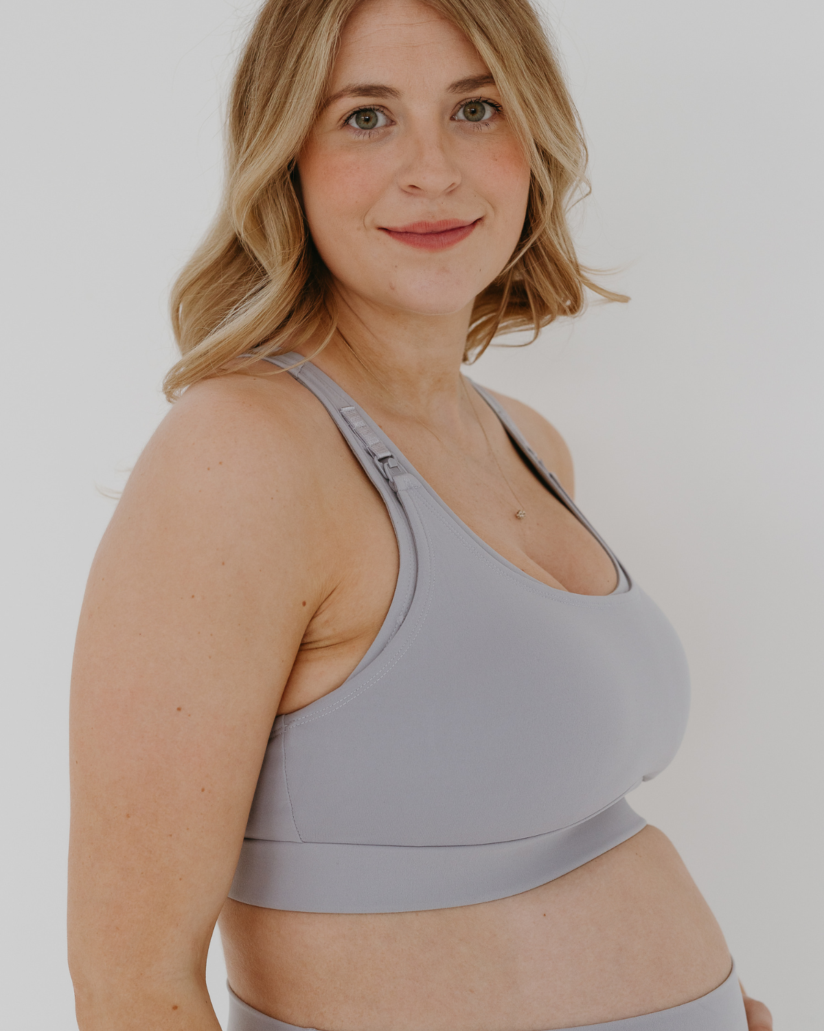 Side view of a pregnant woman wearing a light-gray, high-impact, racerback maternity and nursing bra with clip-down straps, standing against a white background. AXK premium maternity activewear