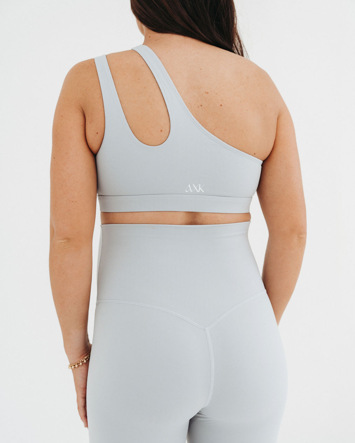 Back view of a pregnant woman wearing a premium light-gray one-shoulder, low-impact maternity and nursing sports bra with clip-down access, paired with high-waist maternity leggings, standing against a light background. Premium maternity athletic and activewear by AXK.