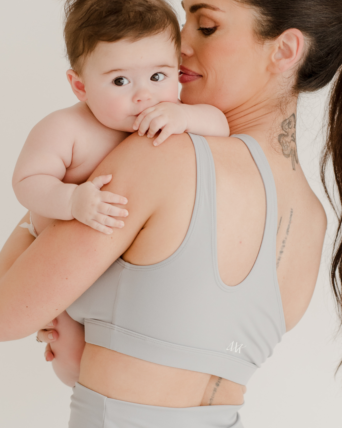Back view of a mother holding baby while wearing a light-gray one-shoulder, nursing sports bra standing in front of a white background. Premium maternity activewear by AXK.