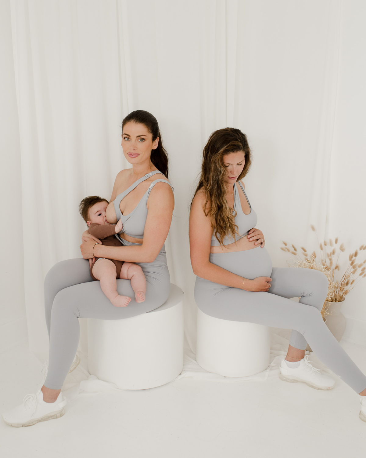 Two women wearing premium light grey maternity leggings with supportive high waists and matching one-shoulder, low-impact, maternity and nursing sports bras, seated against a white background. One is nursing her baby, while the other cradles her pregnant belly. Premium maternity athletic and activewear by AXK.