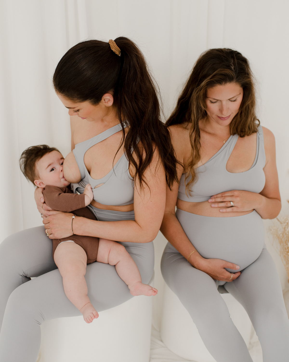 Two women wearing premium light grey maternity leggings with supportive high waists and matching one-shoulder, low-impact, maternity and nursing sports bras, seated against a white background. One is nursing her baby, while the other cradles her pregnant belly. Premium maternity athletic and activewear by AXK.