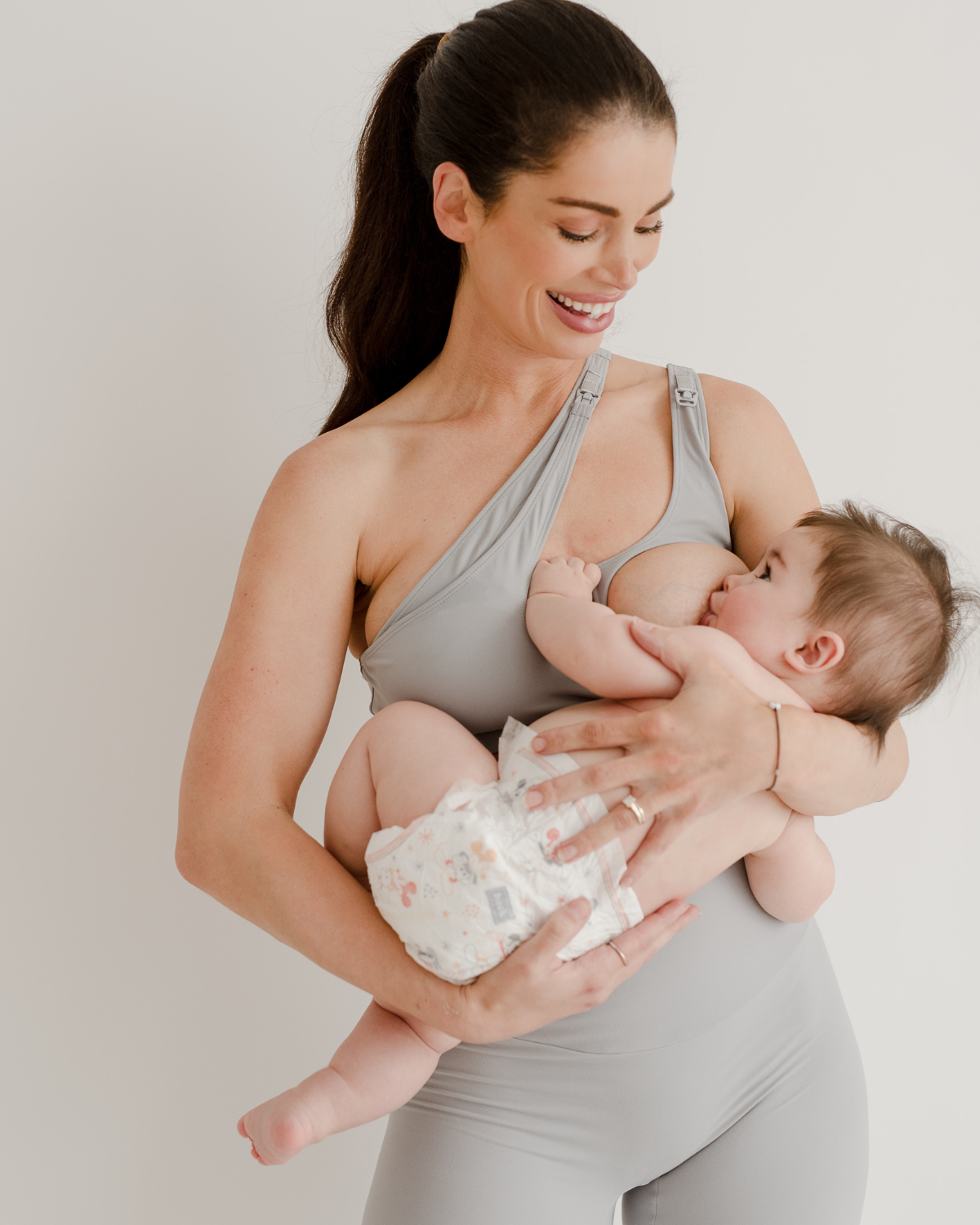 A smiling mother in a light-gray one-shoulder maternity and nursing sports bra and matching leggings, breastfeeding her baby against a white background. The bra features one-handed clip access for easy nursing. Premium maternity activewear by AXK.