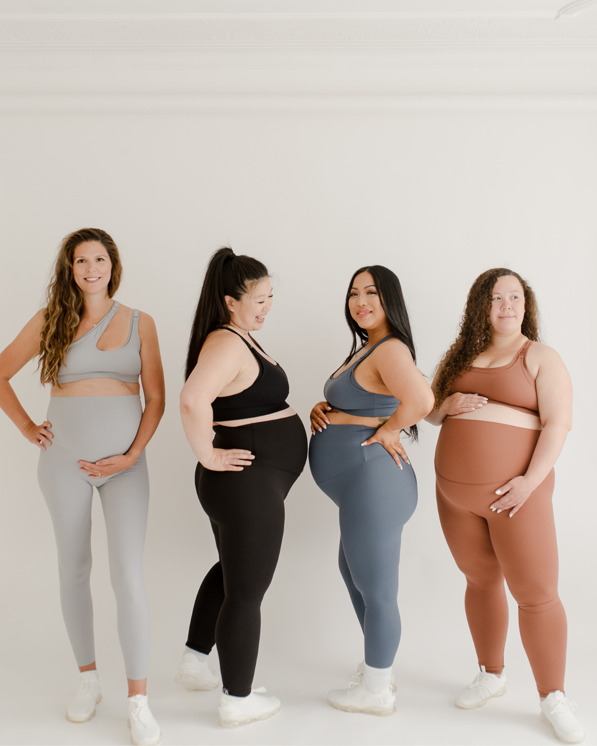 Four pregnant women of diverse body types wearing premium maternity leggings with flattering V-seam backs and matching high-impact, maternity and nursing sports bras in light grey, black, blue, and brown. All standing against a white background. Premium maternity athletic and activewear by AXK.