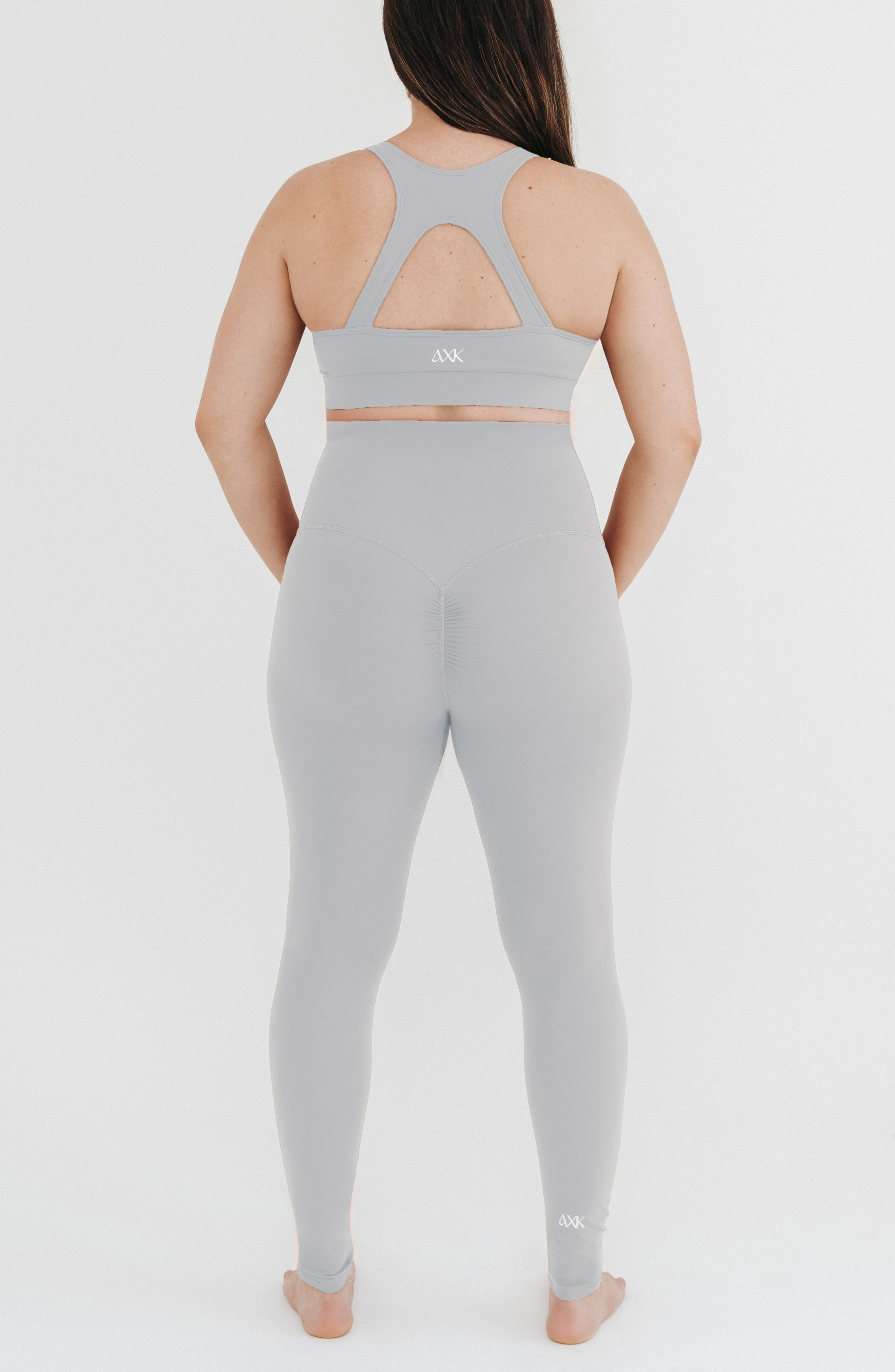 Pregnant woman wearing premium light grey maternity leggings with a flattering scrunch butt and a high impact, racer back nursing sports bra on a white background. Premium maternity athletic and activewear by AXK.