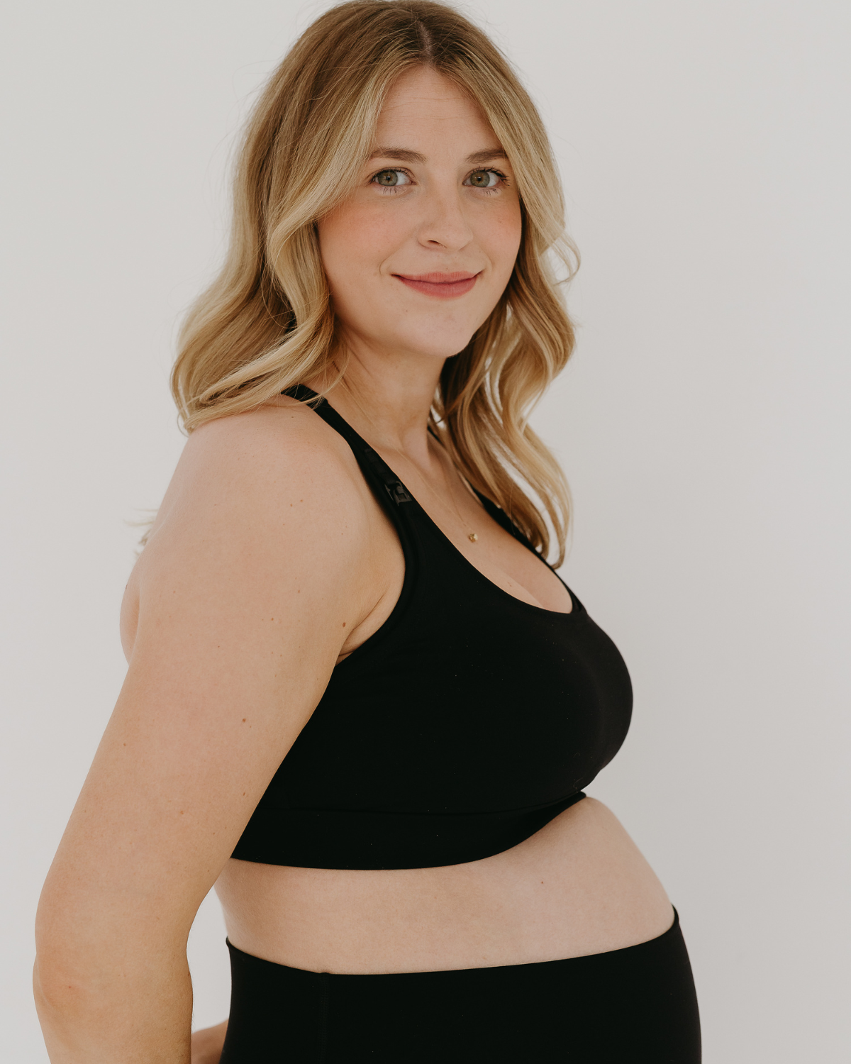 Side view of a pregnant woman wearing a black high-impact, racerback, maternity and nursing sports bra paired with matching high-waist leggings. Premium maternity athletic and activewear by AXK.