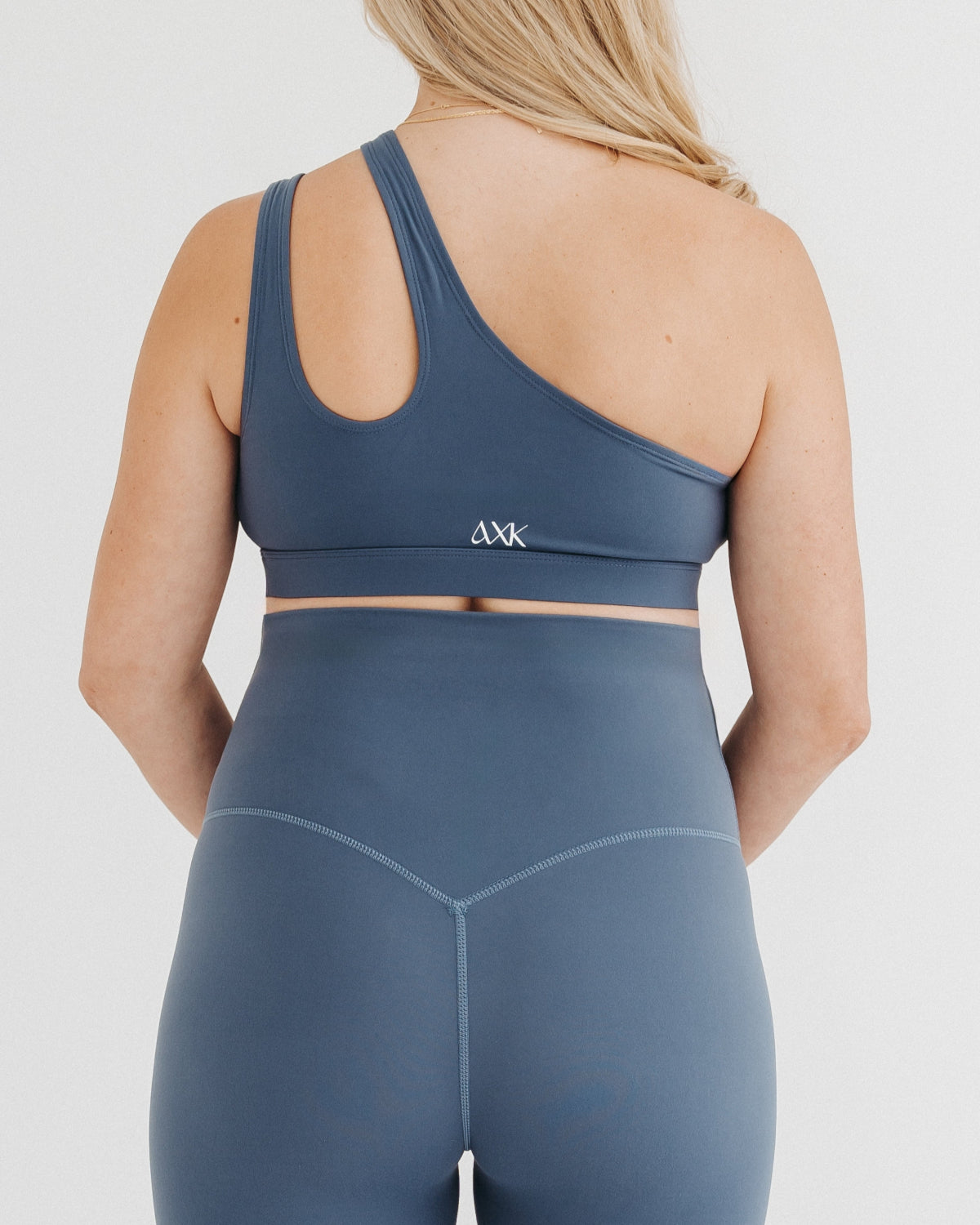 Pregnant woman wearing premium one-shoulder maternity and nursing sports bra in blue. Paired with blue AXK maternity leggings for a stylish maternity activewear set. Premium maternity athletic and athleisure workout clothes by AXK in plus size, tall friendly, and petite friendly options. 