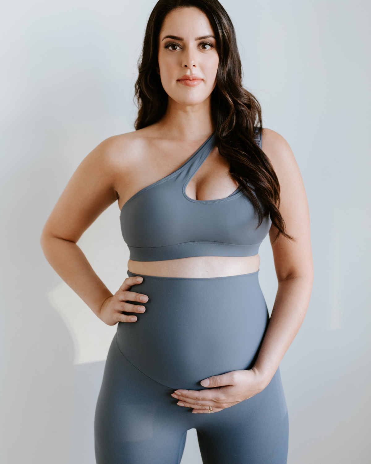 Pregnant woman wearing a blue one-shoulder, maternity, nursing sports bra and matching high-waisted maternity leggings, standing against a white background. Premium maternity activewear by AXK.
