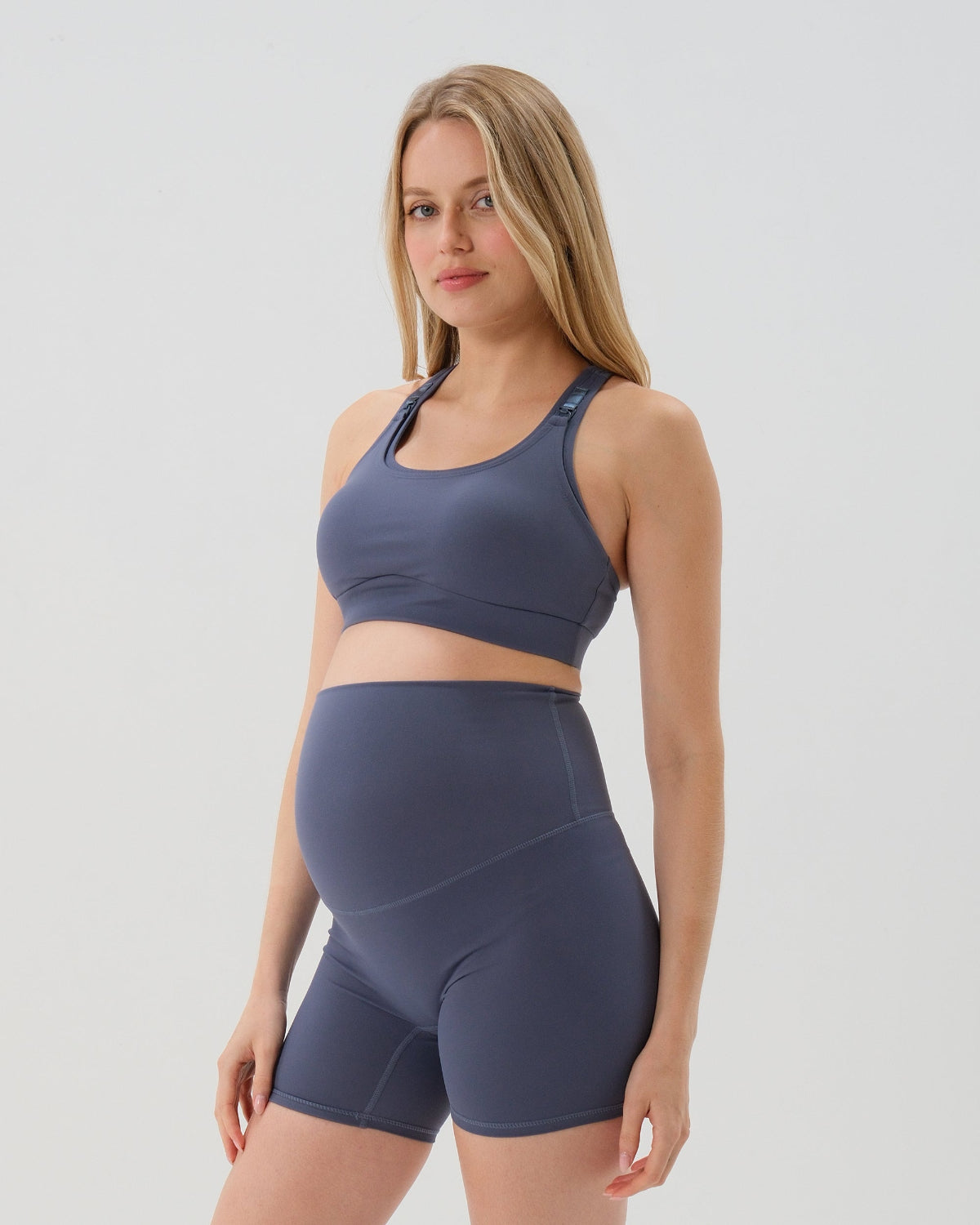 Pregnant woman wearing our premium blue best reviewed maternity bike shorts  for 2026 with a supportive high waist and matching high-impact, racerback maternity and nursing sports bra, standing against a white background. Premium maternity athletic and activewear by AXK.