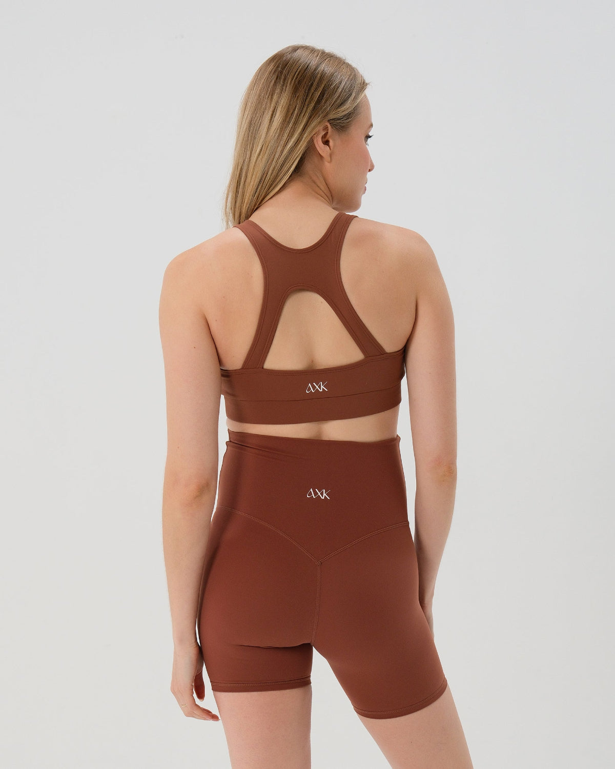 Back view of pregnant woman wearing our best reviewed premium brown maternity bike shorts with a supportive high waist and matching high-impact, racerback maternity and nursing sports bra, standing against a white background. Premium maternity athletic and activewear by AXK - high quality petite, tall, and plus size maternity. 