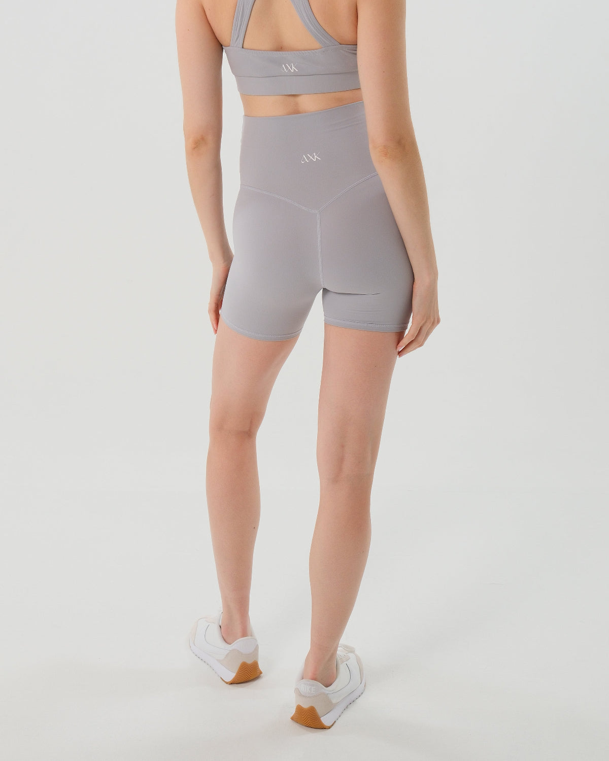 Pregnant woman wearing premium light grey maternity bike shorts with a supportive high waist and matching high-impact, racerback maternity and nursing sports bra, standing against a white background. Premium maternity athletic and activewear by AXK.