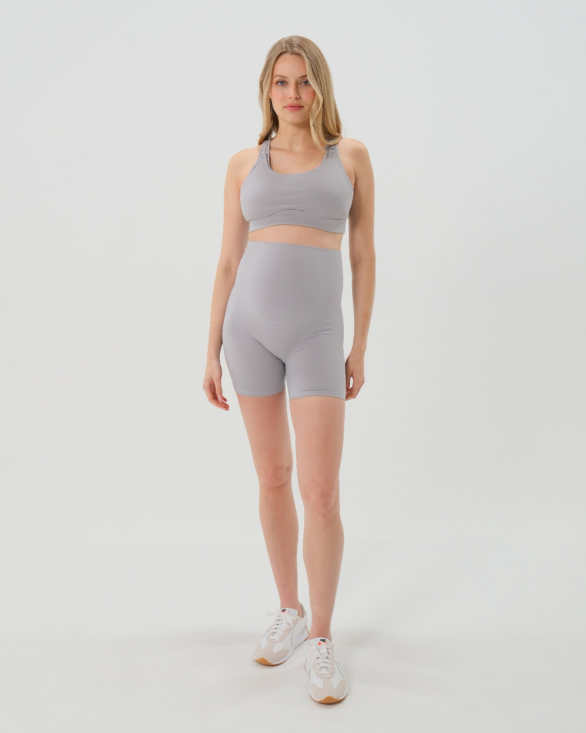 Pregnant woman wearing premium light grey maternity bike shorts with a supportive high waist and matching high-impact, racerback maternity and nursing sports bra, standing against a white background. Premium maternity athletic and activewear by AXK.
