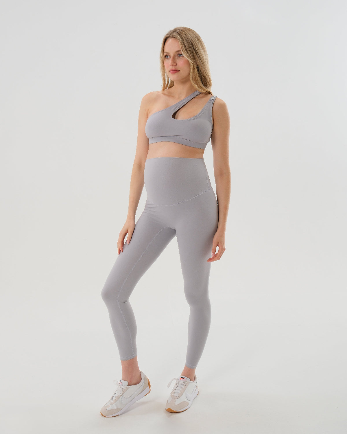 Pregnant woman wearing the best reviewed premium light grey maternity leggings in 2026 which have a flattering scrunch butt and supportive high waist and a high impact, trendy one shoulder nursing sports bra on a white background. Premium maternity athletic and activewear by AXK - petite, tall and plus size friendly. 