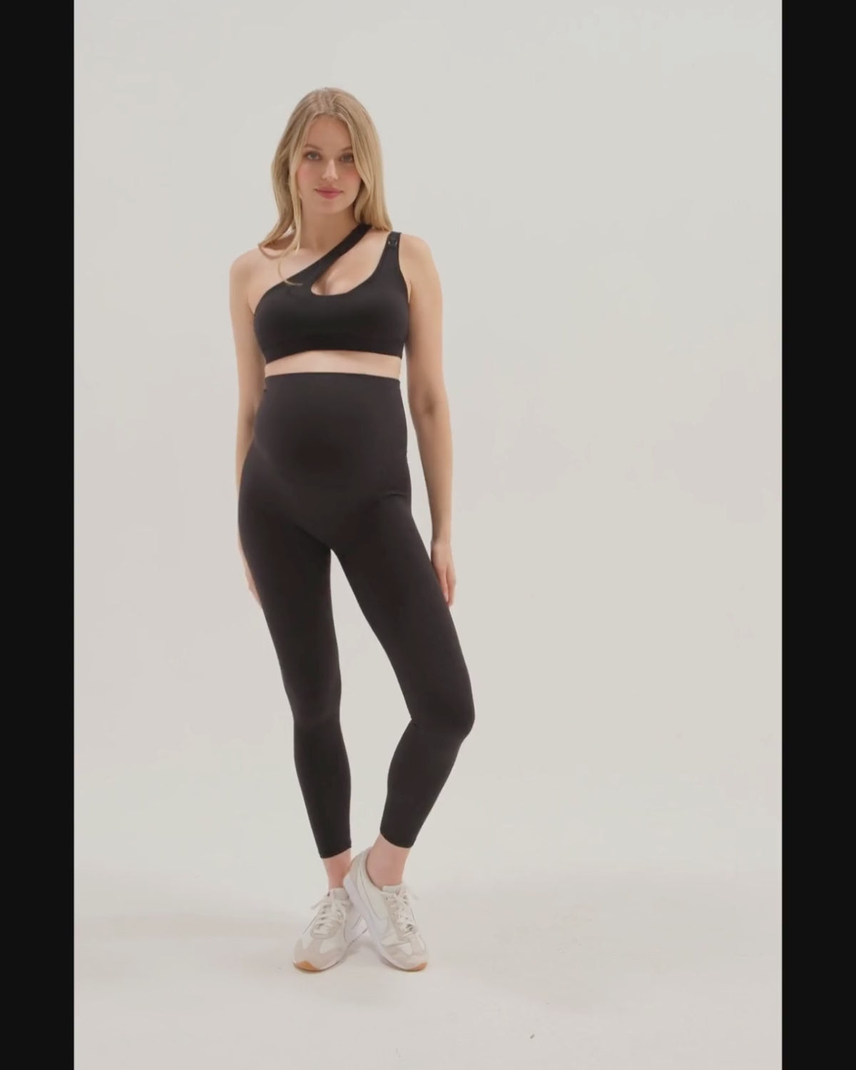 Pregnant woman in a one-shoulder black maternity and nursing sports bra, wearing matching black maternity leggings. Best selling 2026 premium maternity athletic and activewear by AXK, offered in regular and plus sizes, with glowing 5 star reviews. 